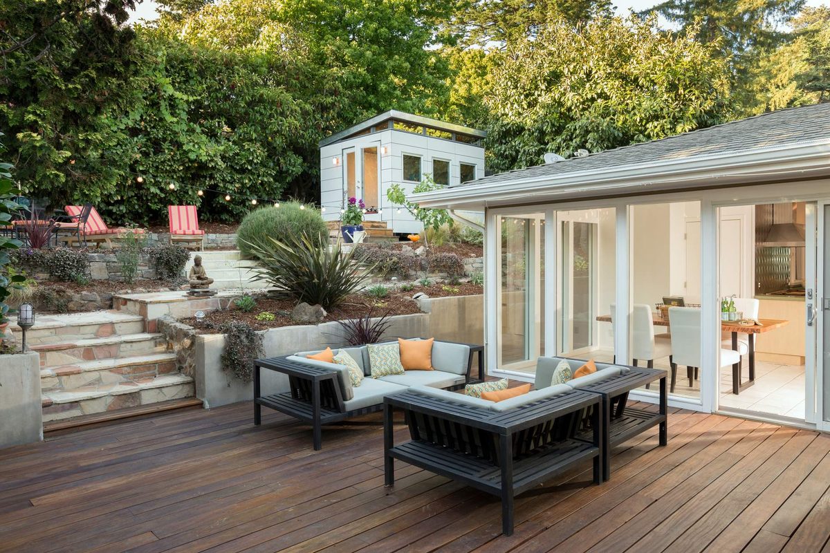 A timber deck with outdoor sofas beside a house with glass doors, garden steps and a small cabin among trees behind