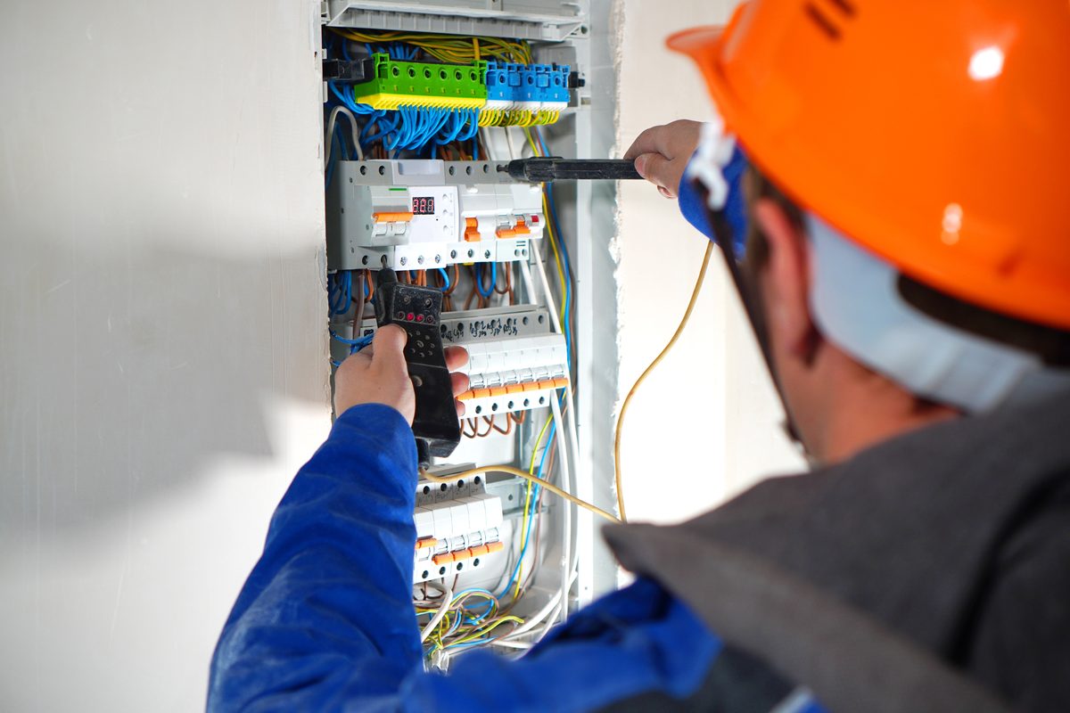 An electrician in an orange hard hat testing circuits in an open switchboard with a multimeter