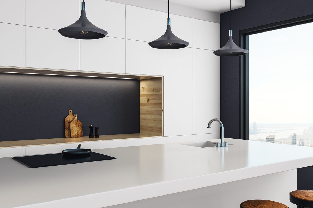 A white kitchen with three black pendant lights over the island bench, a strip of under-cabinet lighting and a city view through a tall window