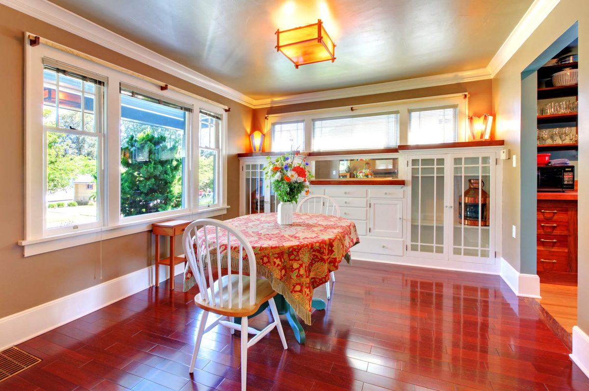 A dining room with tall sash windows in a bay, a lantern-style ceiling light, a round table with a floral cloth and a polished timber floor