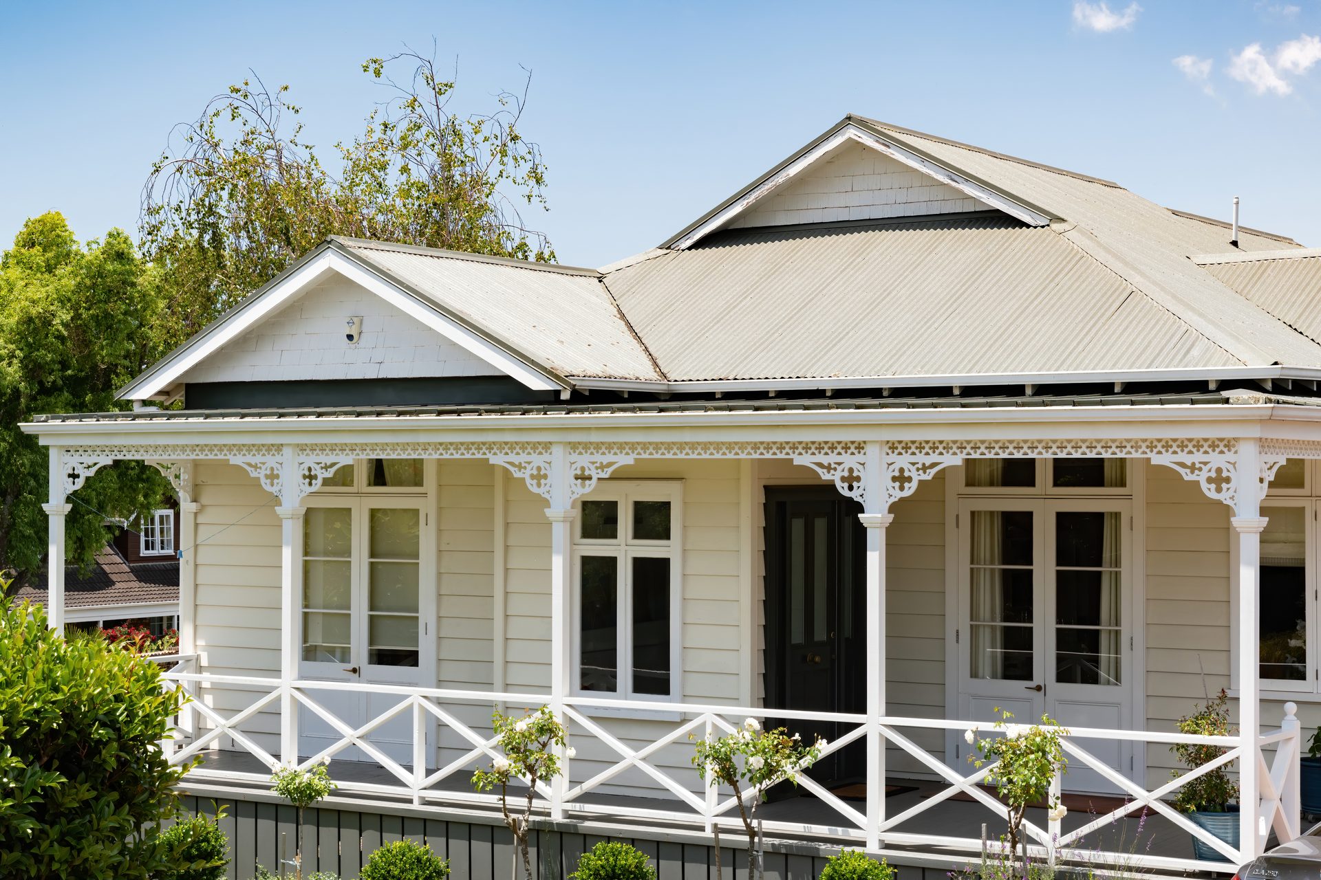 A cream weatherboard villa with a corrugated iron roof, white fretwork brackets along the verandah, French doors and a cross-braced balustrade